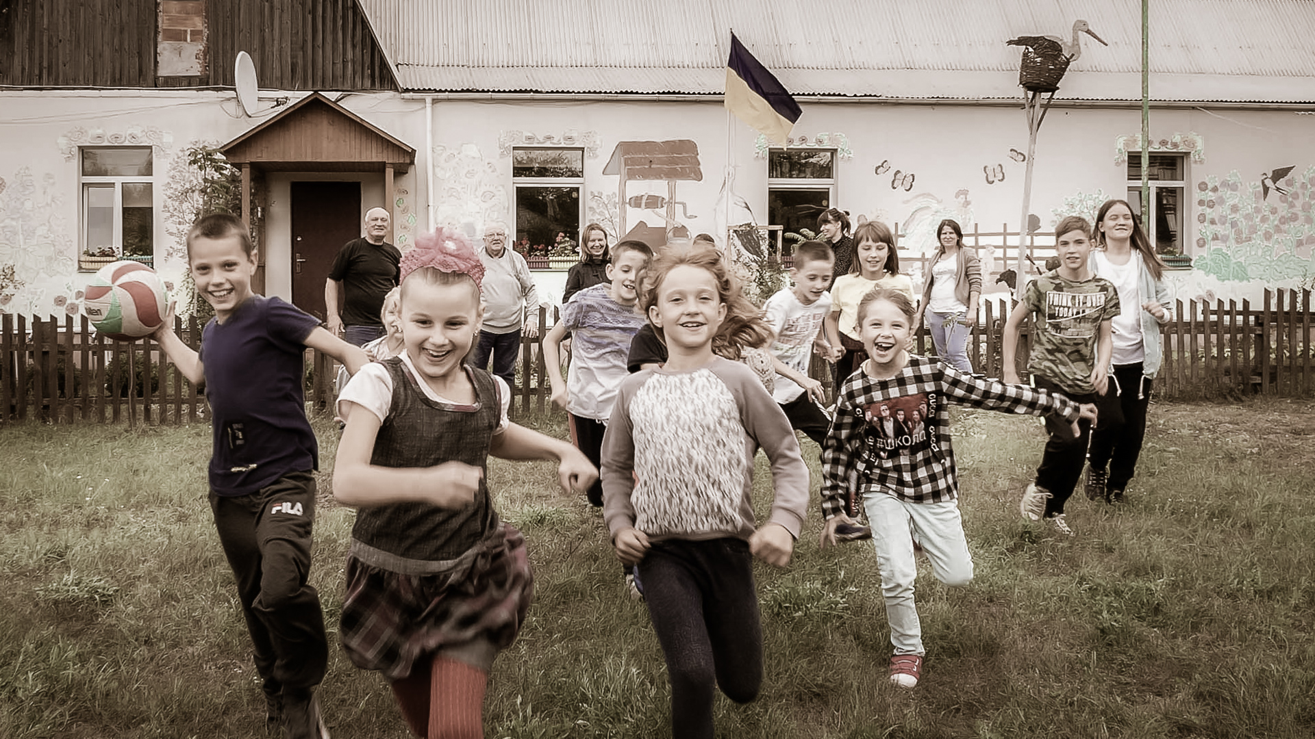 Lapset juoksevat iloisesti pihalla ukrainalaisen Kaverikodin -päiväkeskuksen edessä. Taustalla näkyy talo, jonka seinissä on piirroksia ja Ukrainan lippu.